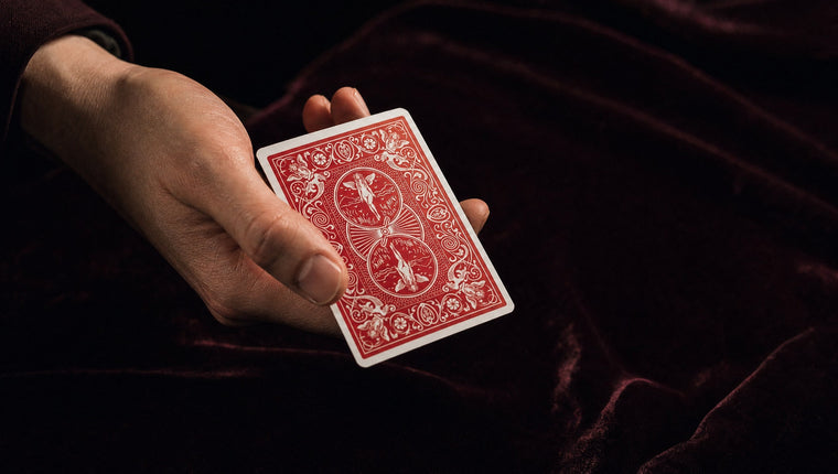A hand holding a single red-backed playing card at an angle over dark velvet