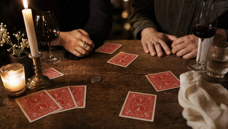 Guests’ hands around a candlelit dinner table scattered with red-backed cards, wine glasses and a coin
